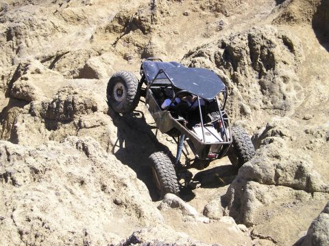 1989 Ford Bronco - Elevated view of a tube-chassis rock buggy with a black flat roof panel, exposed full cage and coilover suspension articulating over pale t…