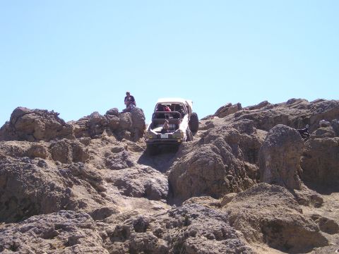 1989 Ford Bronco - Low front-angle shot of a stripped white 4x4 with a doorless caged cab cresting a steep tan rock outcrop against a clear blue sky, front ti…