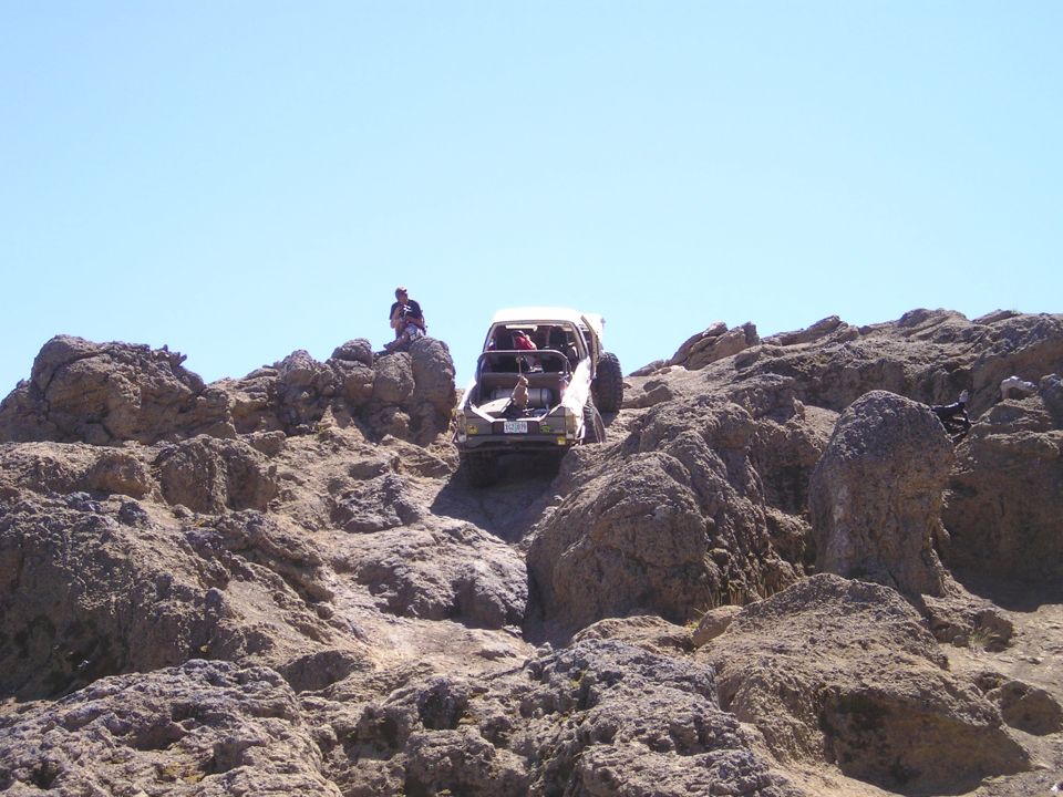 Stripped white caged 4x4 cresting a steep tan rock outcrop with a spectator watching
