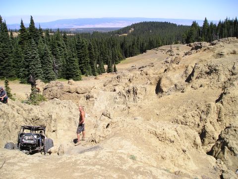 1989 Ford Bronco - Wide view across a tan rock obstacle in an alpine clearing, with a caged rock buggy on oversized mud tires parked at lower left and a shirt…
