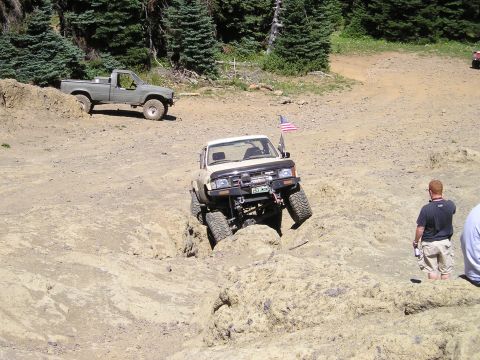 1989 Ford Bronco - Tan lifted mini-truck with a winch-equipped tube front bumper, roof light bar and American flag on a whip drops its front end into a rutted…