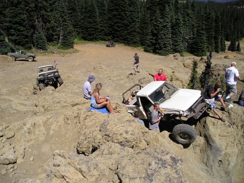 1989 Ford Bronco - Elevated view of a stripped white doorless 4x4 pickup with a roll cage tipped nose-down into a tan rock crevice while a crowd of spectators…