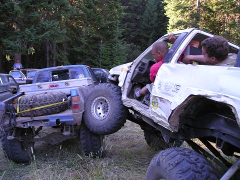 1989 Ford Bronco - Two off-road trucks appear to be stuck together during an event in a wooded area.
