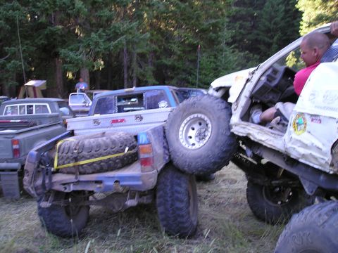 1989 Ford Bronco - Off-road trucks are stuck together in a wooded area with a spare tire on the lower truck's bed.