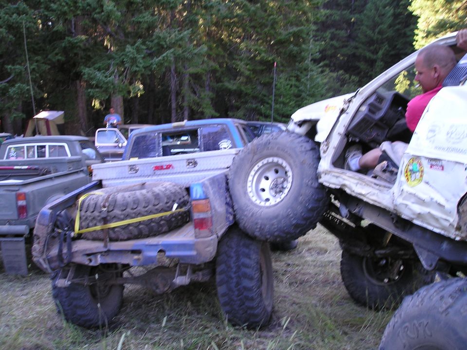 Two off-road trucks are stuck together, one on top of the other, in a wooded area. A spare tire is strapped to the bed of the lower truck