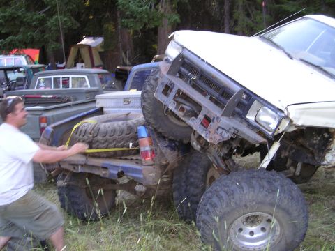 1989 Ford Bronco - A man secures a spare tire to a blue truck bed while a white truck is stuck on top of it.