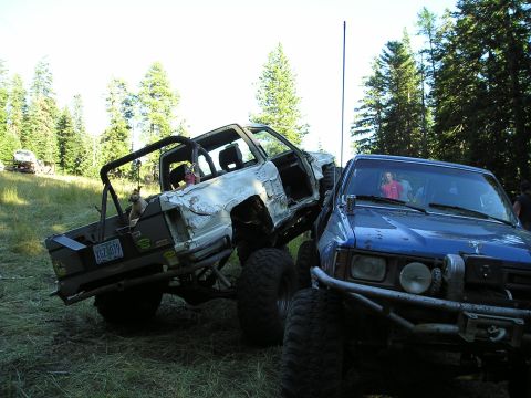 1989 Ford Bronco - Two custom off-road trucks are parked on grassy terrain with trees in the background.