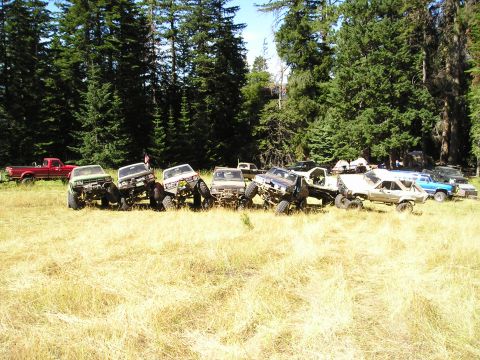 1989 Ford Bronco - A collection of off-road vehicles are parked in a field, with a dense forest behind them.