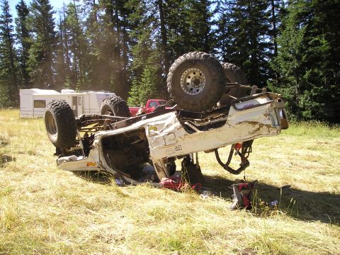 1989 Ford Bronco - A white 1989 Ford Bronco lies overturned in a grassy area, with campers and trees visible behind it.