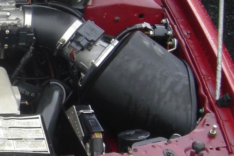 1991 Ford Taurus - Engine-bay close-up of the intake tract in a red car, showing a black plastic airbox connected by a ribbed rubber tube and T-bolt clamps to…