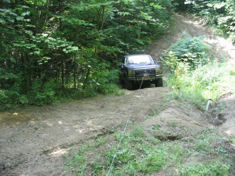 1986 Ford Bronco - A dark-colored Ford Bronco is winched up a muddy incline in a wooded area.