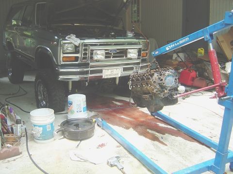 1986 Ford Bronco - An engine is being removed from a lifted Ford Bronco in a garage setting with tools and buckets around.