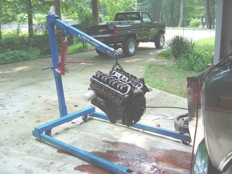 1986 Ford Bronco - A V8 engine is suspended by a blue engine hoist, with a green pickup truck parked nearby.