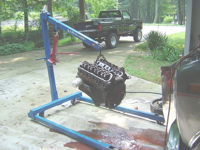 V8 engine hanging from blue hoist on driveway, valve covers off, lifted truck behind