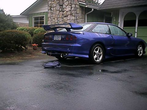 1993 Ford Probe - A blue 1993 Ford Probe is viewed from the rear on a wet driveway, showing its distinctive spoiler and dual exhaust.