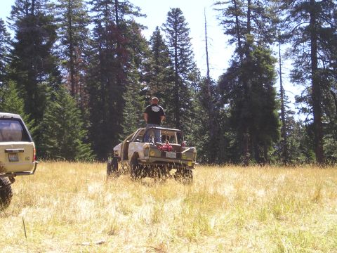 1989 Ford Bronco - Two off-road vehicles are in a field, one with a man standing on its roof.