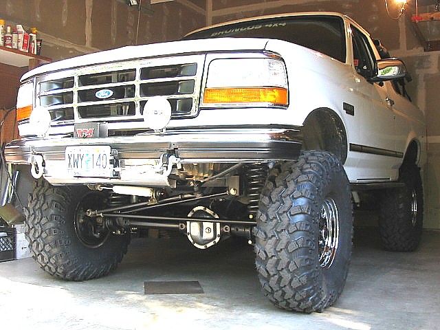 White lifted Ford Bronco on large mud tires in garage, low front 3/4 view showing front axle