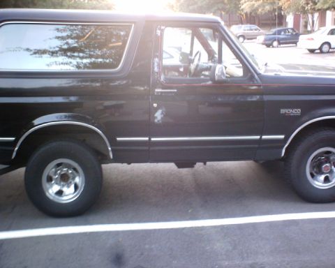 1989 Ford Bronco - The side profile of a black Ford Bronco is visible, parked in a lot with other vehicles in the background.