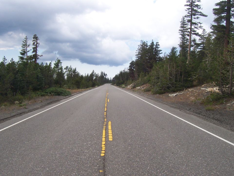 Asphalt road with double yellow lines and white shoulder lines, surrounded by trees under a cloudy sky