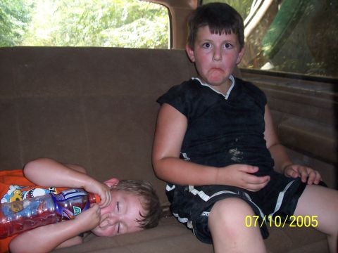 1987 Ford Bronco - Two boys are in the back seat of a vehicle. One boy is lying down drinking from a bottle, while the other sits with a displeased expression.