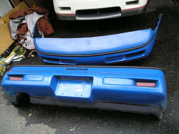 Blue Pontiac bumper with red reflectors and black trim, labeled 'Pontiac'. Another bumper and part of a white car are visible