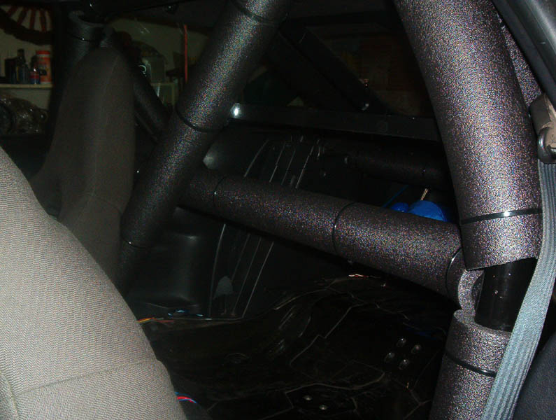 Black textured roll cage with diagonal bracing installed in a stripped rear seat area of a car
