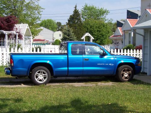 1971 Ford Mustang - A blue pickup truck with 'DAKOTA 5.9 R/T' badging is parked on a grassy lawn in front of a white picket fence and houses.
