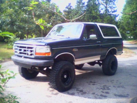 1993 Ford Bronco - Three-quarter front view of a lifted dark green Ford Bronco with tan lower two-tone and a white removable hardtop, parked on a paved drive…