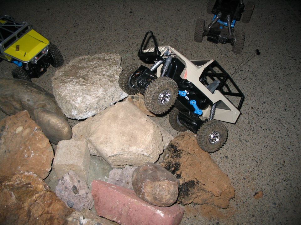 Tan-bodied RC rock crawler with black cage on a pile of rocks, more crawlers nearby at night