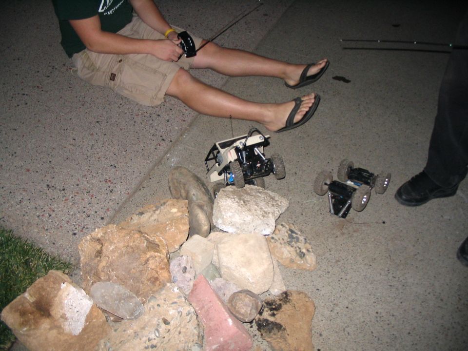 Person in green shirt and shorts sitting holding RC transmitter with two RC rigs on rocks at night