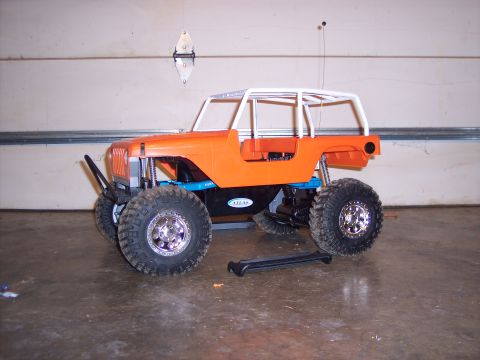 1988 Ford Bronco - An orange RC crawler with a white roll cage and large off-road tires is parked indoors.