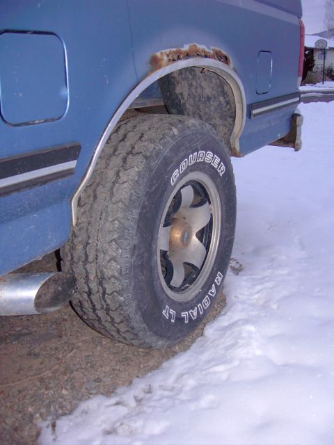 1992 Ford F-150 - A blue pickup truck's rear wheel well shows significant rust and a Courser Radial LT tire, with a chrome exhaust pipe visible.