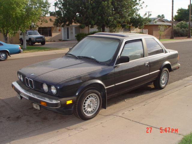 Dark BMW E30 coupe parked on street, white SUV in background