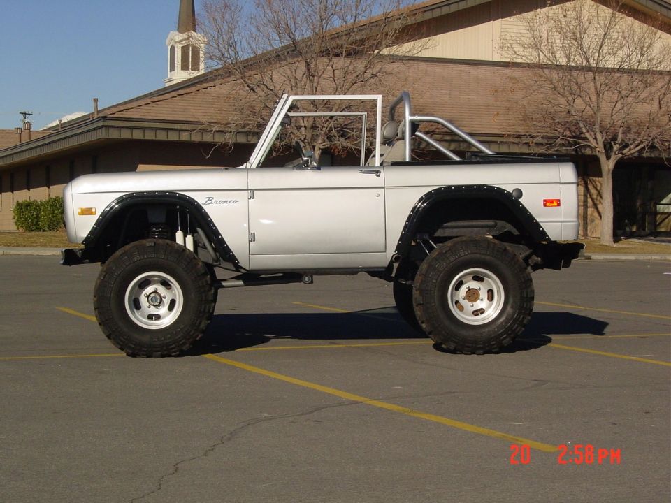 Silver lifted Ford Bronco, topless with roll bar, big mud tires, parked in lot