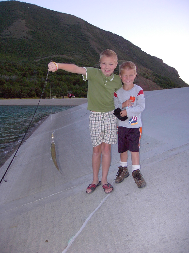 Two young boys stand on a concrete surface, one holding up a small fish on a fishing line. A body of water and a tree-covered hill are in the background