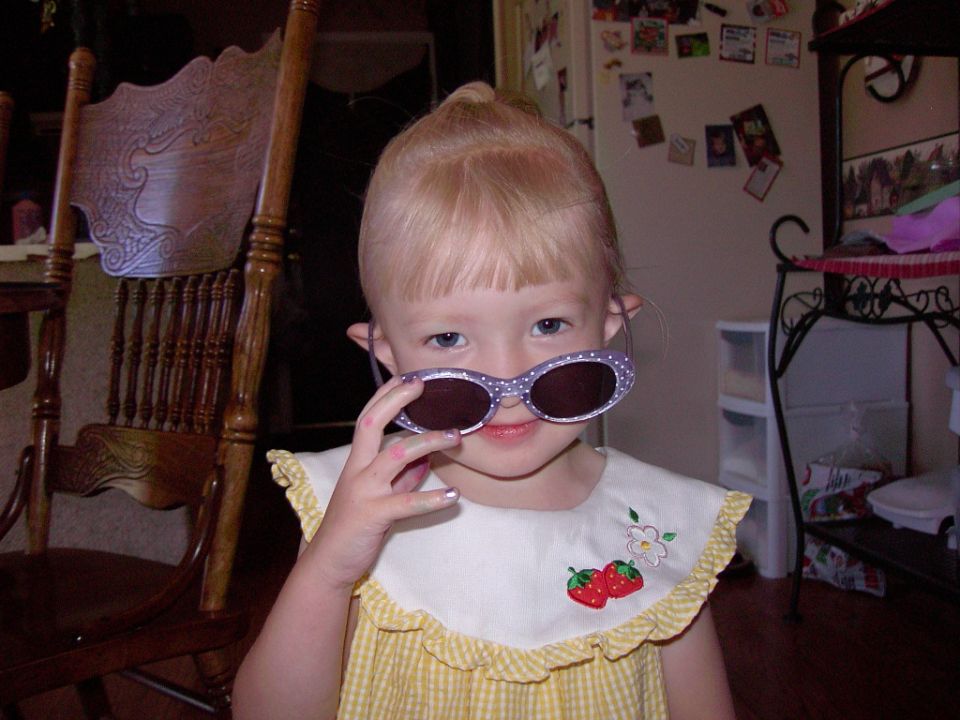 Child with blonde hair and bangs holds purple polka-dot sunglasses to her face