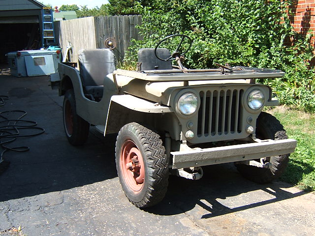 Tan flat-fender Willys Jeep, front 3/4 view, red wheels, on driveway by fence