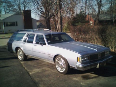 1998 Ford Taurus - A silver station wagon is parked on a paved surface in front of a residential area with trees and houses.