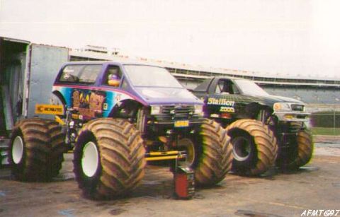 1991 Ford Bronco - Two monster trucks, a purple van-bodied truck and a black pickup-bodied truck, are parked side-by-side.