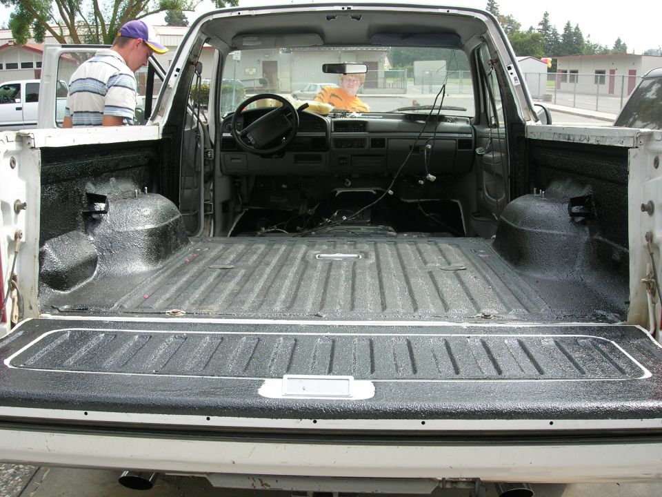 Rear view into white Bronco with gutted interior and fresh black bedliner on floor, wheel wells, and open tailgate