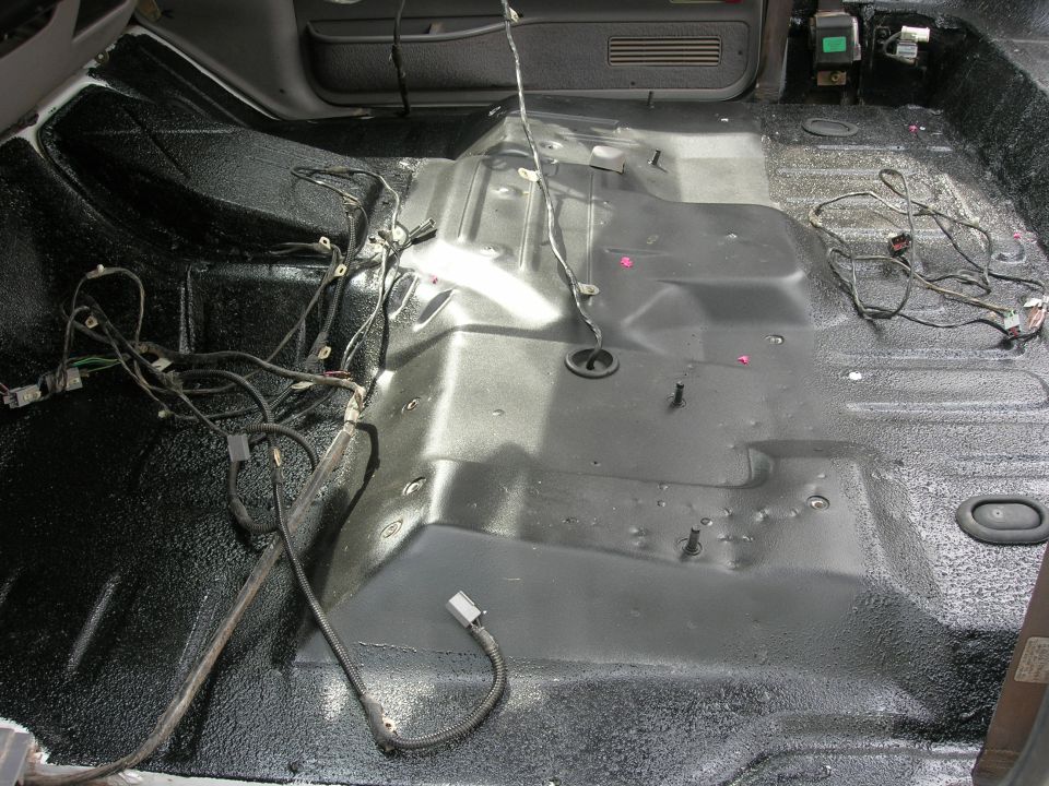 vehicle interior with floor stripped bare, showing wiring and black spray-on bedliner material