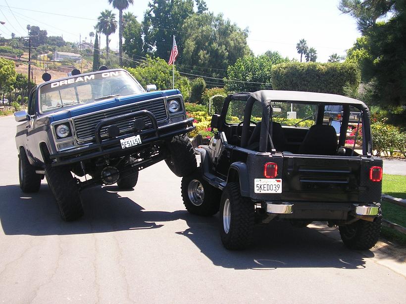 Blue pickup truck with "DREAM ON" on windshield is lifting its front wheels off the ground, with a black Jeep behind it
