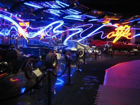 1968 Ford Mustang - Vintage vehicles are displayed in a museum exhibit illuminated by vibrant neon lighting.