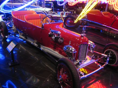 1968 Ford Mustang - A vintage red race car with an open cockpit is displayed indoors, surrounded by colorful neon lighting.