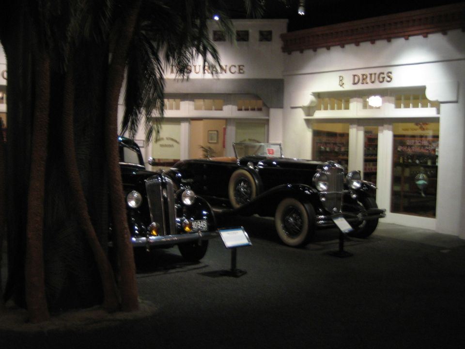 Two old black cars parked in front of a building facade with 'Insurance' and 'Drugs' signs