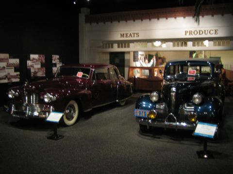 1968 Ford Mustang - A burgundy sedan and a dark blue sedan are displayed in a museum exhibit, with a backdrop simulating a storefront.