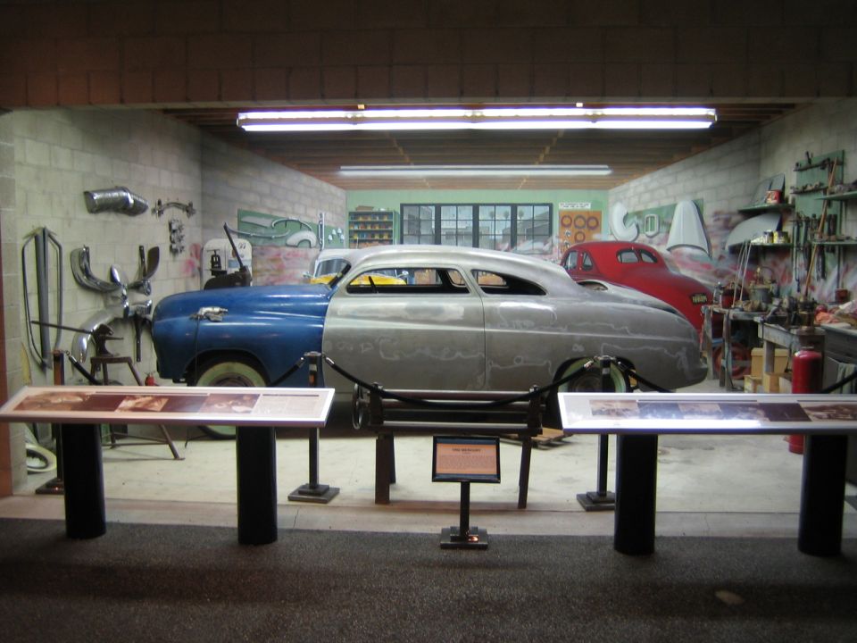 Car in a workshop, half blue and half bare metal, with a red car in the background