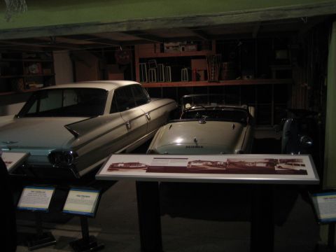 1968 Ford Mustang - A white sedan and a convertible are displayed indoors, with informational signs in front of them.