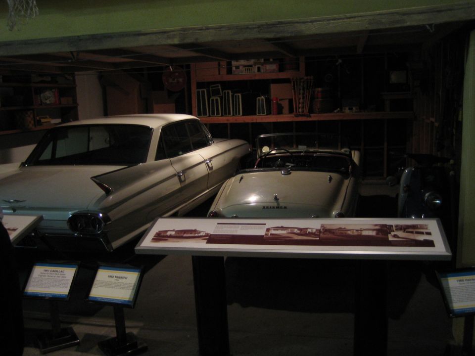 Two vintage cars, a white sedan and a light-colored convertible, parked indoors with display signs