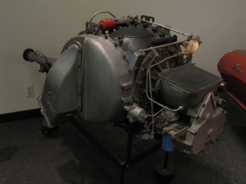 1968 Ford Mustang - A complex aircraft engine is displayed on a stand, with a portion of a red vehicle in the background.